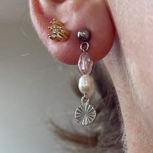 May include: Close-up of an ear with two earrings. One is a gold-coloured stud with a hand design. The other is a dangling earring with a silver stud, pink bead, white pearl, and a silver sunburst charm.