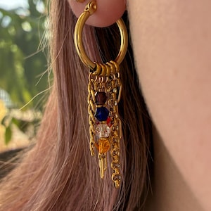 Peut inclure: Boucles d'oreilles créoles dorées avec des chaînes pendantes et des perles colorées. Les boucles d'oreilles comportent une petite boucle d'oreille à clous dorée avec un groupe de petits cristaux. Les créoles ont une série de petites chaînes avec des perles rouges, bleues et oranges.