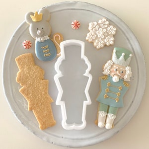 May include: A set of five Christmas-themed cookie cutters, including a nutcracker, a snowflake, a mouse, and two other shapes. The cutters are made of white plastic and are arranged on a white plate.