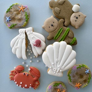 Puede incluir: Una colección de galletas de azúcar decoradas con una variedad de diseños con temática marina, incluyendo conchas marinas, un pulpo, un cangrejo y nutrias marinas. Las galletas están decoradas con glaseado blanco, marrón, rojo y verde.
