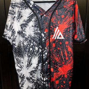 May include: A black baseball jersey with a white and red splatter paint design. The jersey has a button-up front and a black collar. The left side of the jersey features white splatter paint, while the right side features red splatter paint. A white triangle with a black outline and a black letter "A" inside is on the right side of the jersey.