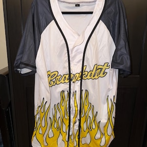 May include: White baseball jersey with black sleeves and trim. The jersey features yellow flames at the bottom and the word "Brawkout" in yellow script across the chest. The jersey is hanging on a black hanger.