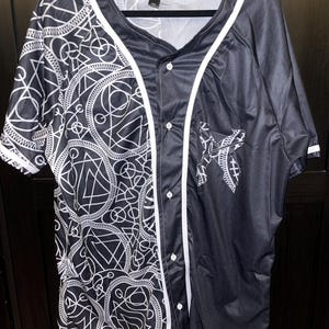 May include: A black and white baseball jersey with a geometric pattern on the left side and a star design on the right side. The jersey has a button-up front and white piping.