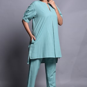 May include: A solid light teal two-piece set consisting of a tunic and trousers. The tunic features a split neckline, short sleeves with cuffs, and a central vertical pleat. The matching trousers are straight-legged and ankle-length.