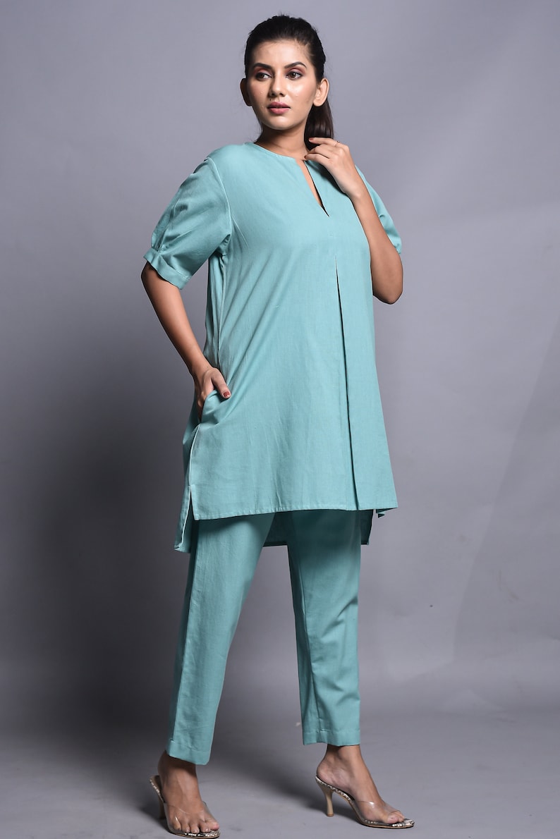May include: A solid light teal two-piece set consisting of a tunic and trousers. The tunic features a split neckline, short sleeves with cuffs, and a central vertical pleat. The matching trousers are straight-legged and ankle-length.
