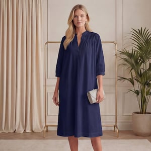 May include: A navy blue midi dress with a V-neck and three-quarter sleeves. The dress has a relaxed fit and falls to the mid-calf. A small, light-colored clutch is held in the hand. The dress is displayed in a well-lit room.