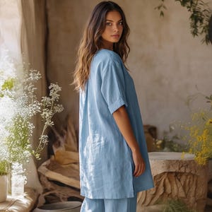 May include: A light blue tunic top with short sleeves and a side slit, featuring a relaxed fit and a smooth, solid-colored fabric. The garment has a simple round neckline and falls to the mid-thigh, paired with matching light blue trousers.