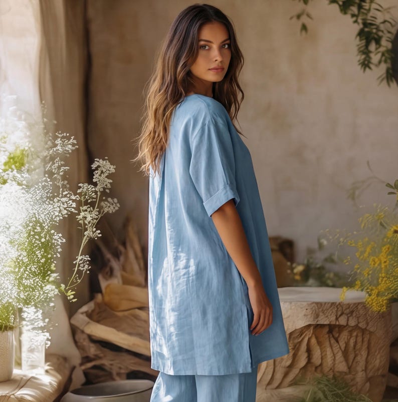 May include: A light blue tunic top with short sleeves and a side slit, featuring a relaxed fit and a smooth, solid-colored fabric. The garment has a simple round neckline and falls to the mid-thigh, paired with matching light blue trousers.
