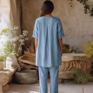 May include: A light blue matching set consisting of a short-sleeved tunic top with a central back pleat and cuffed sleeves, paired with matching straight-leg trousers. The garment features a solid color and a relaxed, loose-fitting silhouette.