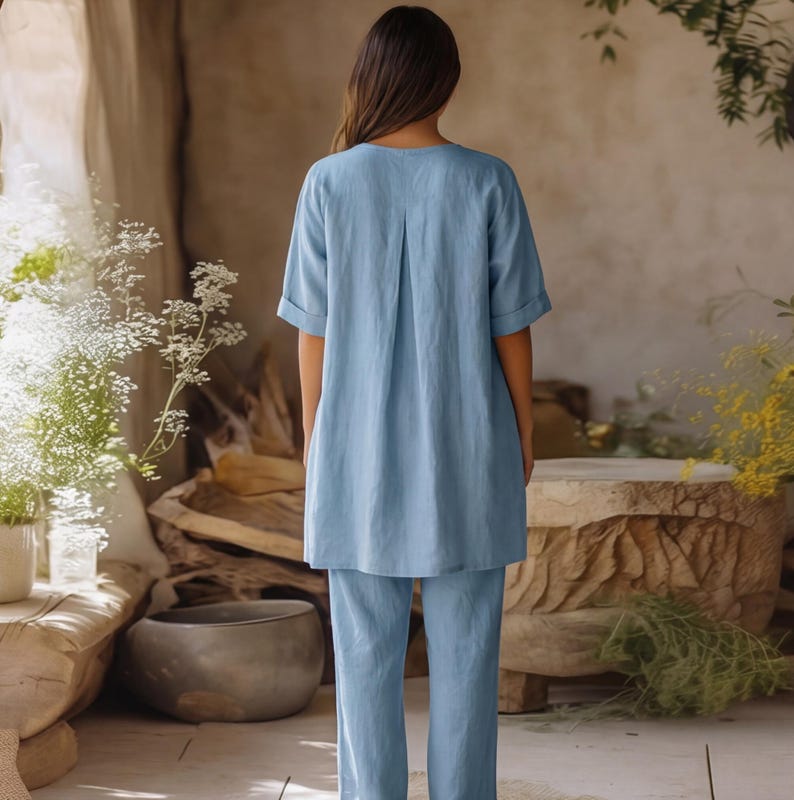 May include: A light blue matching set consisting of a short-sleeved tunic top with a central back pleat and cuffed sleeves, paired with matching straight-leg trousers. The garment features a solid color and a relaxed, loose-fitting silhouette.
