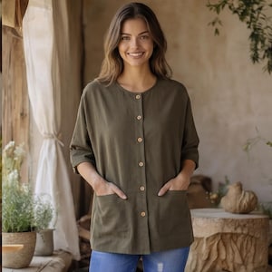May include: Olive green button-down shirt with a round neckline and two front pockets. The shirt has three-quarter sleeves and wooden buttons. The model is wearing blue jeans. The shirt is made of a soft, comfortable material.