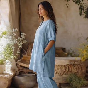 May include: A light blue tunic top with a V-neckline, short sleeves, and side pockets, paired with matching light blue trousers. The garment features a straight hemline with side slits, providing a relaxed fit and a solid, uniform color throughout.