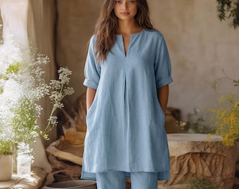May include: A light blue tunic top with a V-neckline, short cuffed sleeves, and side pockets, paired with matching light blue trousers. The ensemble features a clean, solid-colored design with a relaxed fit and a straight hemline on the tunic.