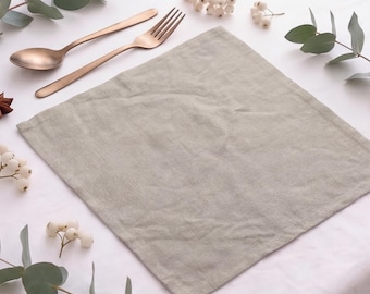 May include: A square, light beige linen napkin placed centrally, accompanied by a copper-toned metal spoon and fork. The arrangement is accented with sprigs of green eucalyptus leaves and small clusters of white berries on a white surface.