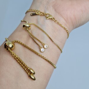 May include: Three gold chain bracelets with adjustable sliding clasps. Two of the bracelets have small clear crystal accents. The bracelets are layered on a person's wrist.