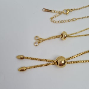 May include: A gold chain necklace with two adjustable lengths. The necklace has a small round pendant with a clear crystal on each end. The chain is made of stainless steel.