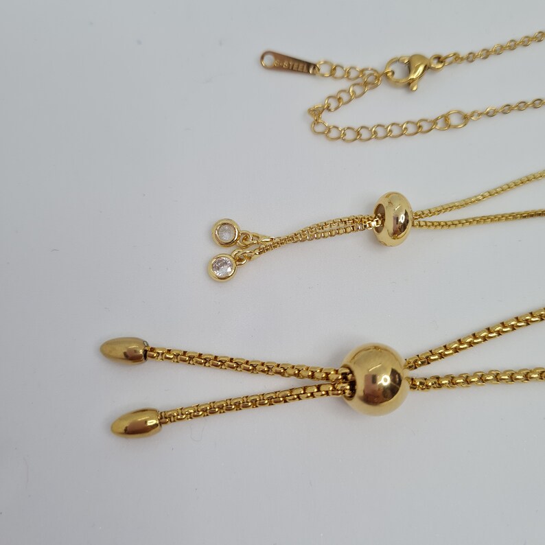 May include: A gold chain necklace with two adjustable lengths. The necklace has a small round pendant with a clear crystal on each end. The chain is made of stainless steel.