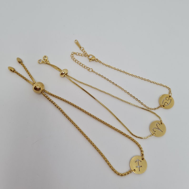 May include: Three gold adjustable bracelets with zodiac signs. The bracelets feature a delicate chain with a small round charm featuring the zodiac signs of Sagittarius, Aries, and Cancer.