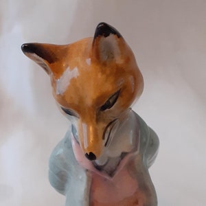 May include: A ceramic figurine of a fox wearing a blue jacket and pink waistcoat. The fox is standing on a green base with brown and pink flowers.
