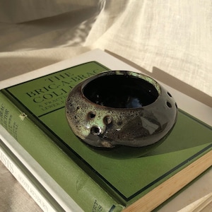 Może przedstawiać: Mała, ciemnozielona i czarna ceramiczna doniczka z otworami po bokach. Doniczka stoi na zielonej książce.