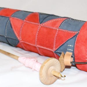 May include: A red and blue patchwork cylinder case with a matching red end cap and white stitching. A wooden spindle with pink yarn is in front of the case. The case is likely for storing yarn or other craft supplies.