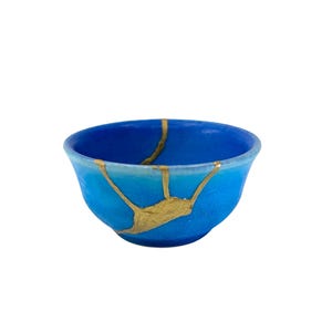 May include: A small, blue ceramic bowl with gold kintsugi repair lines. The bowl features a gradient of blue hues, from a darker shade at the base to a lighter shade near the rim. The gold lines provide a decorative element.
