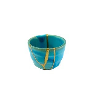 May include: A small, turquoise ceramic cup with gold kintsugi repair lines. The cup has an irregular shape and a smooth, glossy finish. The kintsugi technique highlights the beauty of imperfections. The cup is approximately 7 cm in diameter.