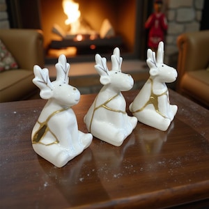 May include: Three white ceramic reindeer figurines with gold accents. The reindeer are sitting with their legs tucked under them. The antlers are detailed with leaf-like shapes. The gold accents appear to be part of a kintsugi design.