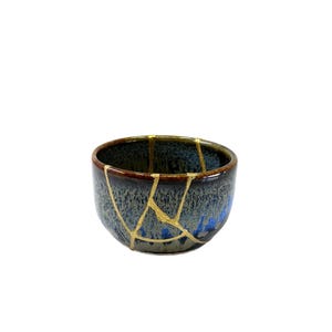 May include: A ceramic bowl with a dark blue and brown glaze, featuring gold-coloured kintsugi repairs. The bowl has a rounded shape and is likely used for serving or display. The kintsugi technique highlights the history of the object.