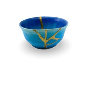 May include: A blue ceramic bowl with gold kintsugi repair lines. The bowl features a gradient of blue shades, lighter at the base and deepening towards the rim. The gold lines accentuate the cracks, a traditional Japanese art form.