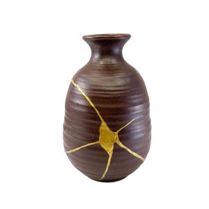 May include: A brown ceramic vase with a narrow neck and a rounded body. The vase features gold-coloured kintsugi, or repaired cracks, adding a decorative element. The vase is approximately 15 cm tall.