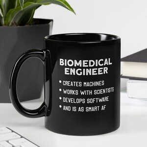 Op de afbeelding: Zwarte keramische mok met witte tekst "BIOMEDICAL ENGINEER" en taakomschrijvingen. De mok zegt ook "AND IS AS SMART AF". De mok staat op een wit oppervlak.
