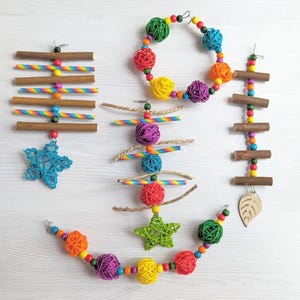Puede incluir: Un conjunto de coloridos juguetes para pájaros. Los juguetes incluyen palos de madera, bolas tejidas y adornos en forma de estrella en varios colores como rojo, verde, amarillo y azul. Algunos juguetes tienen detalles de arcoíris y cuentas de madera.