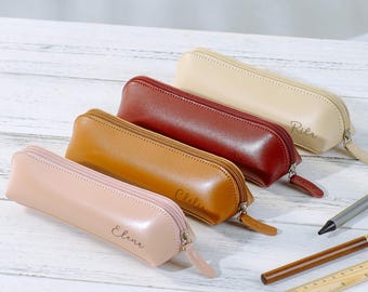 May include: Four leather pencil cases in shades of pink, tan, brown, and burgundy are arranged on a white wooden surface. Each case has a zipper closure and a small tag. The names "Elena", "Chelie", and "Pilar" are inscribed on the cases.