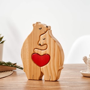 Puede incluir: Escultura de madera de dos osos abraz&aacute;ndose con un coraz&oacute;n rojo en el centro. Los osos est&aacute;n hechos de madera marr&oacute;n claro y tienen rasgos simples y estilizados.