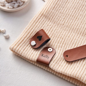 May include: Two brown leather hat clips with a heart and the name "Kelly" engraved on them. The clips are attached to a beige knitted beanie.
