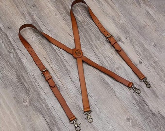 May include: Brown leather suspenders with a metal buckle and a monogrammed leather patch. The suspenders have a Y-shaped back and are adjustable.