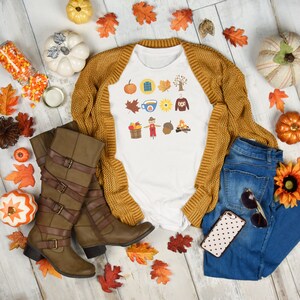 May include: A white t-shirt with a fall-themed design featuring a cup of coffee, a scarecrow, a sunflower, and a campfire. The shirt is paired with a brown cardigan, blue jeans, brown boots, and autumn-coloured leaves.