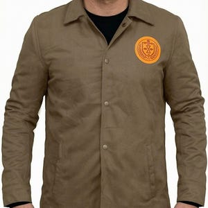 May include: A tan button-down jacket with a classic collar and long sleeves. The jacket features a circular orange patch on the left chest displaying a graphic design of a wheat wreath surrounding a central geometric symbol with horizontal lines.
