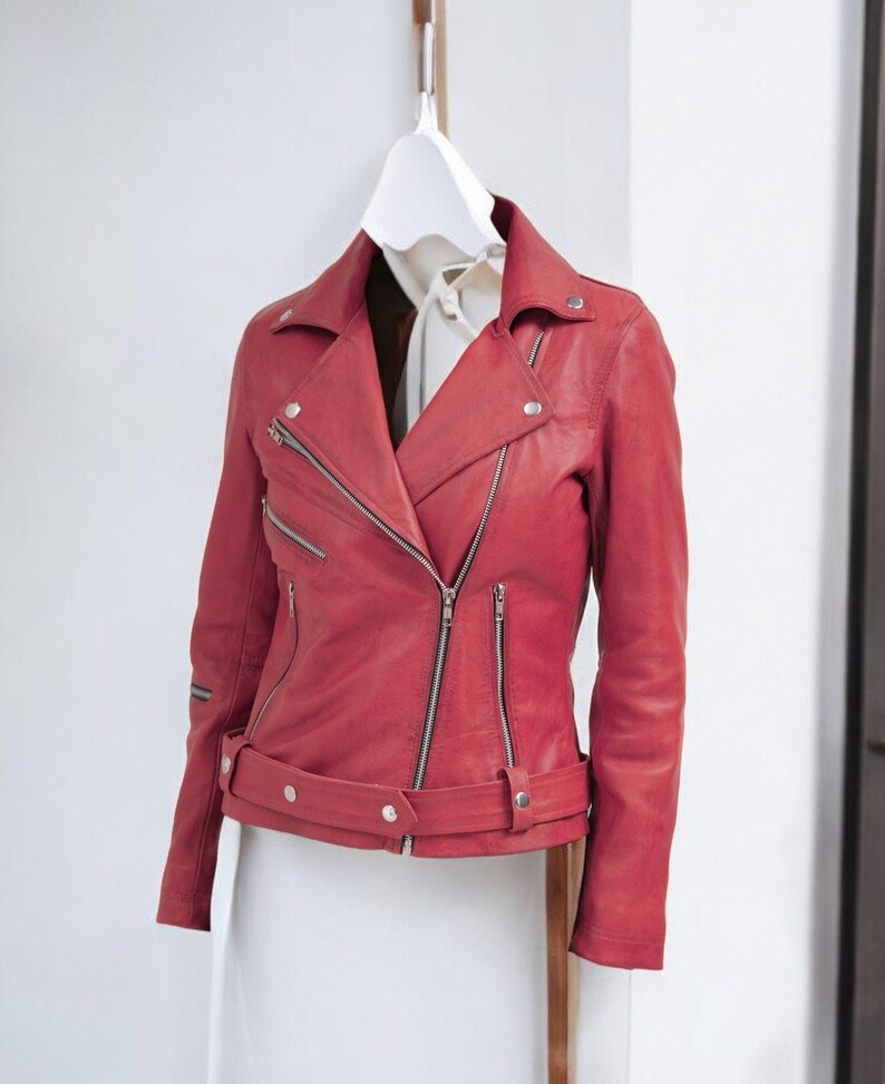 Puede incluir: Chaqueta de cuero roja de estilo motero cl&aacute;sico, con cuello de solapa, cierre de cremallera asim&eacute;trico y m&uacute;ltiples bolsillos con cremallera. La chaqueta tiene cintur&oacute;n en la cintura y est&aacute; colgada en una percha blanca.