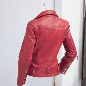 Puede incluir: Chaqueta de cuero roja de estilo motero cl&aacute;sico. La chaqueta presenta un cuello con solapas, un cintur&oacute;n en la cintura y mangas con cremallera. La parte trasera de la chaqueta es visible, mostrando el dise&ntilde;o a medida y los detalles de costura. La chaqueta est&aacute; colgada en una pared blanca.