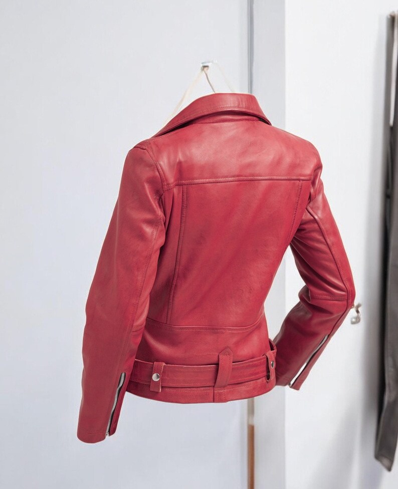 Puede incluir: Chaqueta de cuero roja de estilo motero cl&aacute;sico. La chaqueta presenta un cuello con solapas, un cintur&oacute;n en la cintura y mangas con cremallera. La parte trasera de la chaqueta es visible, mostrando el dise&ntilde;o a medida y los detalles de costura. La chaqueta est&aacute; colgada en una pared blanca.