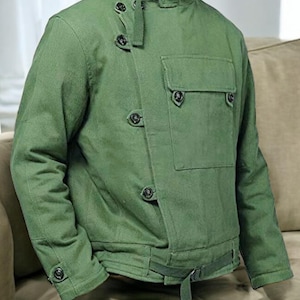 May include: A green jacket featuring a high collar, button fastenings, and a chest pocket. The jacket includes a waist belt and button details on the sleeves. The fabric has a textured appearance.