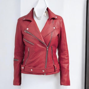 Puede incluir: Chaqueta de cuero roja con un estilo motero cl&aacute;sico. La chaqueta tiene un cuello con solapas, m&uacute;ltiples bolsillos con cremallera y un cintur&oacute;n en la cintura. La chaqueta est&aacute; colgada en una percha blanca contra una pared blanca.