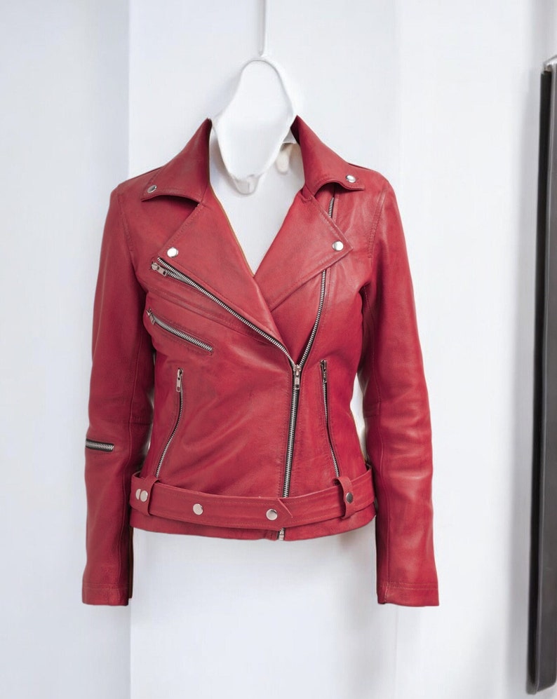 Puede incluir: Chaqueta de cuero roja con un estilo motero cl&aacute;sico. La chaqueta tiene un cuello con solapas, m&uacute;ltiples bolsillos con cremallera y un cintur&oacute;n en la cintura. La chaqueta est&aacute; colgada en una percha blanca contra una pared blanca.