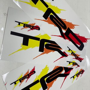 K&ouml;nnte beinhalten: Drei wei&szlig;e Bl&auml;tter Papier mit schwarzen, roten und gelben "TRD" Aufklebern. Die Aufkleber haben ein stilisiertes, Pinselstrich-Design.