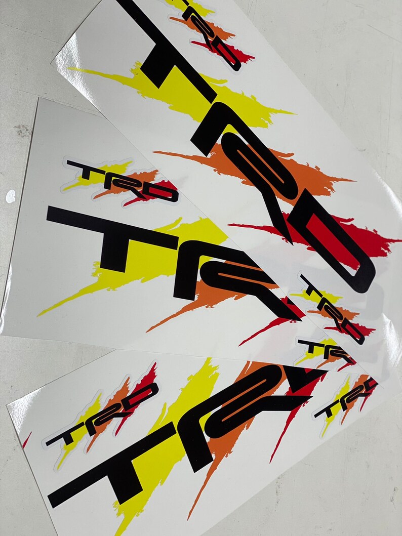K&ouml;nnte beinhalten: Drei wei&szlig;e Bl&auml;tter Papier mit schwarzen, roten und gelben "TRD" Aufklebern. Die Aufkleber haben ein stilisiertes, Pinselstrich-Design.