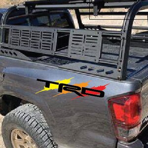 K&ouml;nnte beinhalten: Ein grauer Pickup mit einem schwarzen Metall-Ladepritschen-Rack und einem "TRD"-Aufkleber an der Seite. Der Pickup hat eine schwarze Ladefl&auml;chenverkleidung und eine schwarze Heckklappe.
