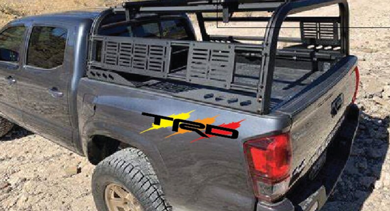 K&ouml;nnte beinhalten: Ein grauer Pickup mit einem schwarzen Metall-Ladepritschen-Rack und einem "TRD"-Aufkleber an der Seite. Der Pickup hat eine schwarze Ladefl&auml;chenverkleidung und eine schwarze Heckklappe.