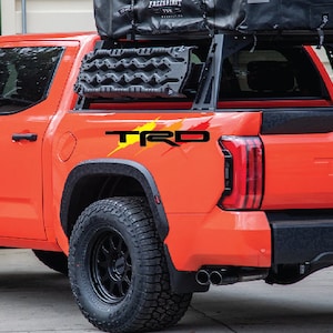 K&ouml;nnte beinhalten: Ein leuchtend orangefarbener Pick-up mit einem schwarzen Dachgep&auml;cktr&auml;ger und einem schwarzen "TRD PRO" Aufkleber auf der Heckklappe. Der Wagen hat schwarze R&auml;der und Reifen.